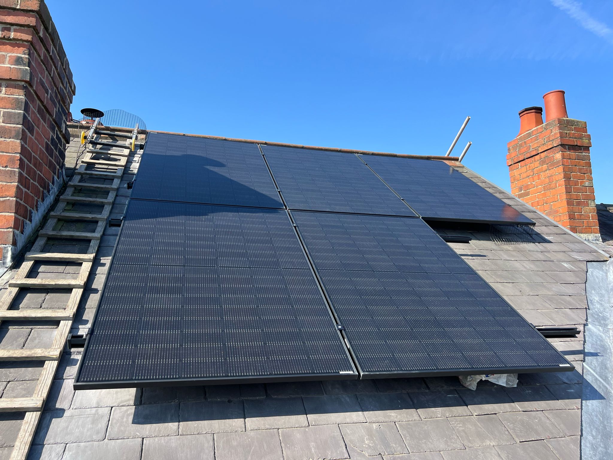 Black solar panels on a slate roof with a ladder and red brick chimneys
